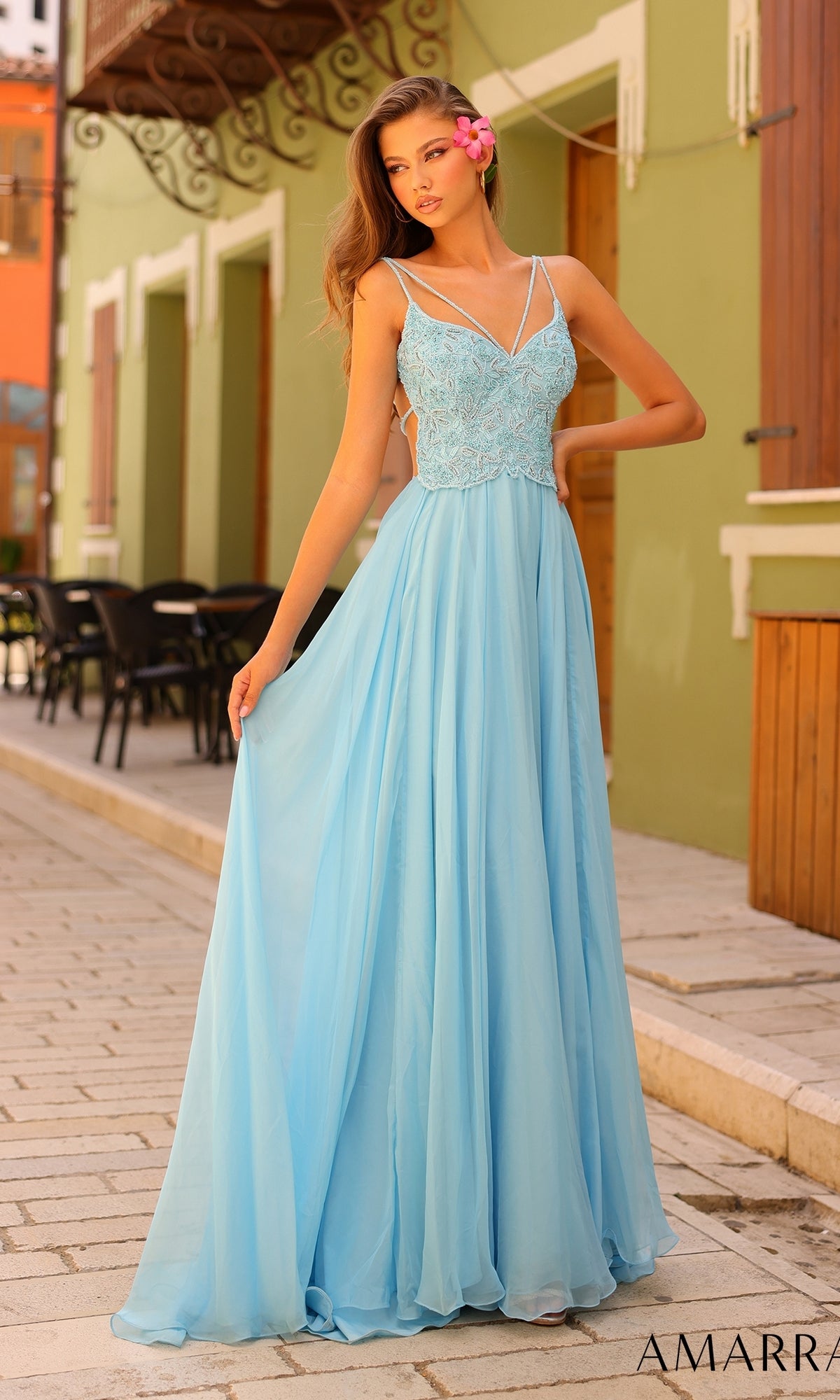 A woman in the Amarra Open-Back Long A-Line Prom Dress 94008, featuring a light blue beaded bodice and spaghetti straps, stands on a cobblestone street, gracefully holding one side of her floor-length gown.